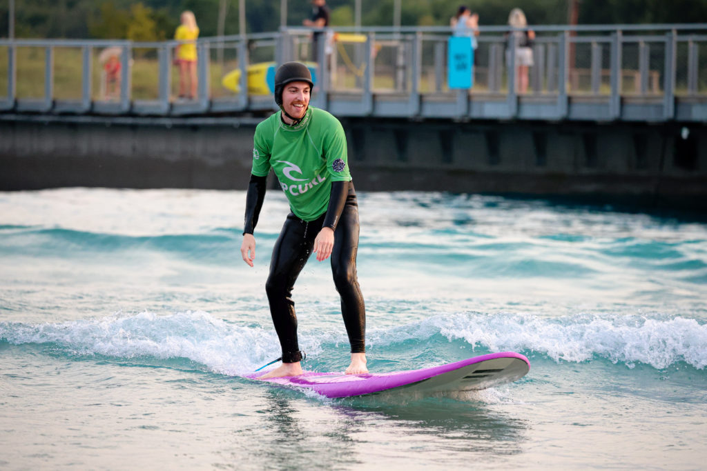 A photo of Kieran, a white man, standing on a surfboard at the wave, an inland surf school